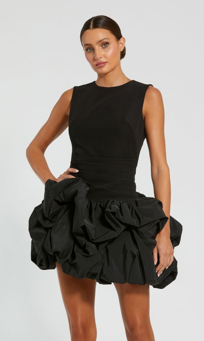 A woman stands against a plain background wearing the Mac Duggal 20709 Short Black Bubble Party Dress, featuring a sleeveless fitted bodice and voluminous mini bubble skirt.